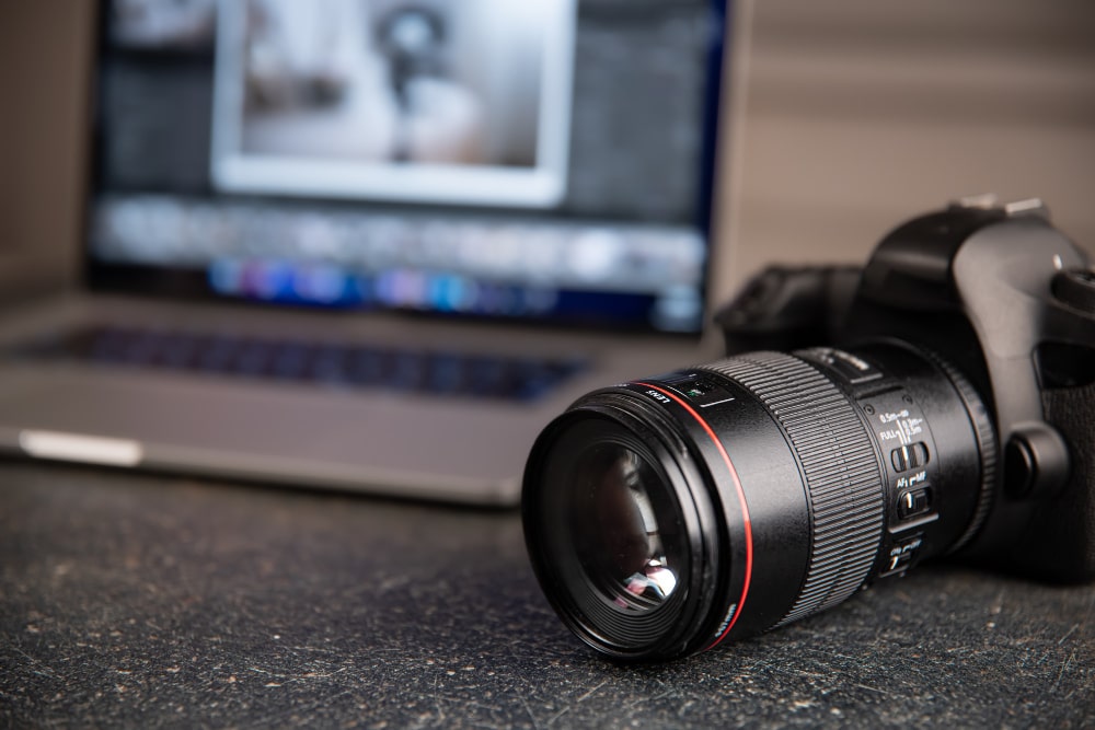 Appareil photo professionnel avec objectif macro à anneau rouge devant un ordinateur portable affichant un logiciel d'édition photo, équipement typique utilisé par les photographes de l'Agence NurPhoto