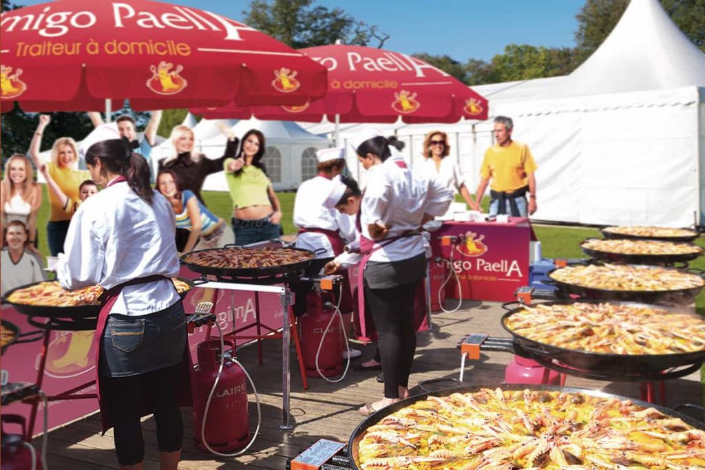 Une paella géante magnifiquement présentée et prête à être servie, avec une pince et des citrons, évoquant un service de "paella géante à domicile" pour des événements