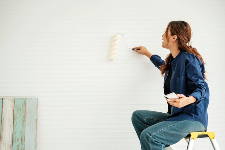 Une femme assise sur un escabeau applique de la peinture blanche avec un rouleau lors d'un projet de interior wall painting.