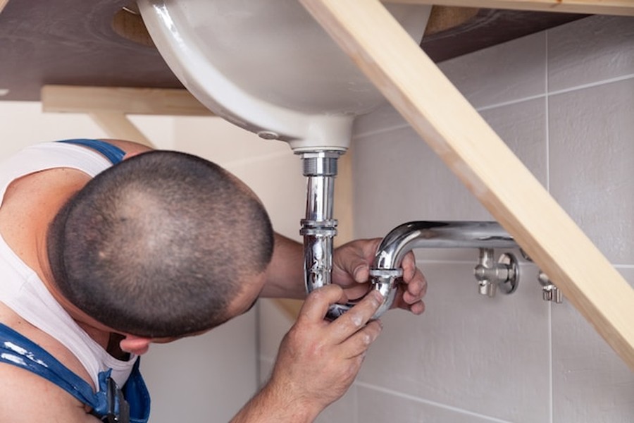 Un plombier professionnel ajuste les raccords chromés d'une plumbing installation sous un lavabo blanc.