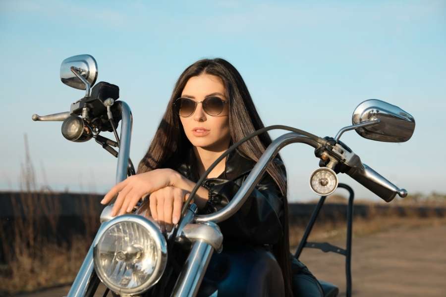 Une jeune femme en veste de cuir noir et lunettes de soleil posant sur une moto custom chromée, incarnant le style et la liberté d'acheter une moto neuve.