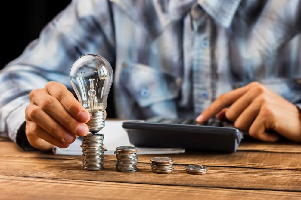 Une personne utilise une calculatrice à côté de piles de pièces de monnaie sur lesquelles repose une ampoule, illustrant comment trouver l'énergie moins chère pour votre entreprise : électricité et gaz.