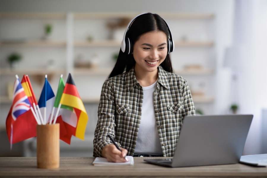 Une étudiante souriante suit un cours de langue en ligne avec un casque et prend des notes, avec des drapeaux internationaux sur son bureau pour surmonter la barrière de la langue.