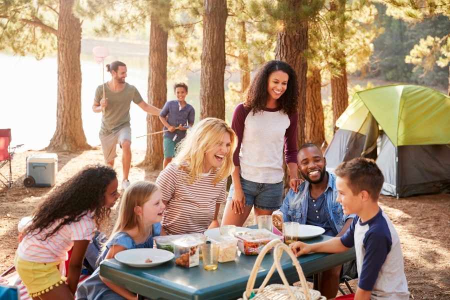 Plusieurs familles profitent d'un premier camping en famille en partageant un repas convivial autour d'une table en bois près d'une tente.