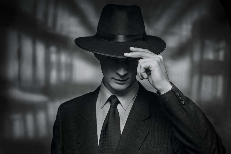 Un homme en costume et chapeau ajustant son fedora, style détective privé.