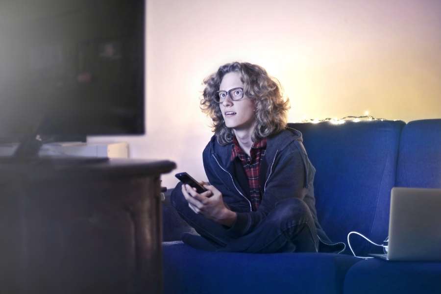 Un jeune homme aux cheveux bouclés portant des lunettes regarde la télévision dans son salon avec un meilleur abonnement IPTV 4K.