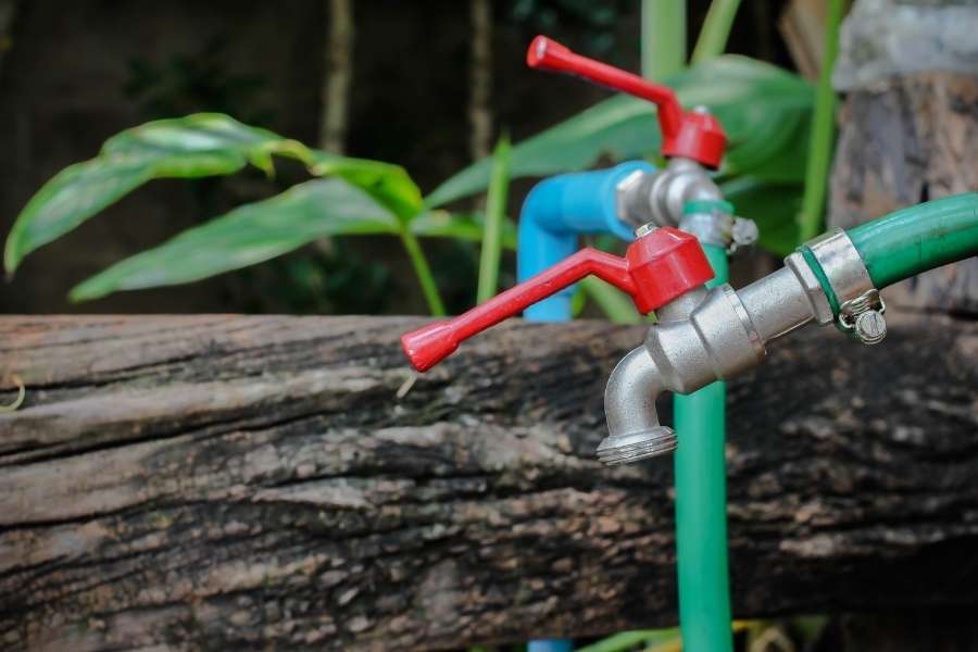 Un robinet de jardin métallique équipé d'une manette rouge et relié à un tuyau vert en extérieur.