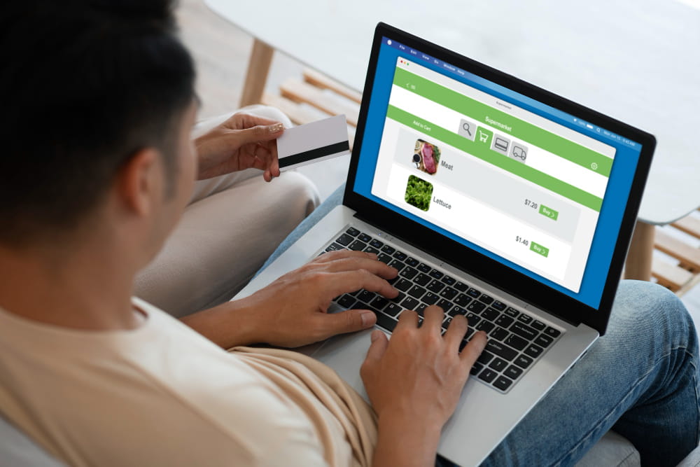 Personne effectuant un achat en ligne de produits d'épicerie sur un ordinateur portable, tenant une carte de crédit. Illustration pour des conseils sur où acheter ou vendre un site internet e-commerce.